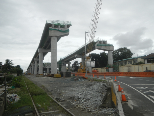 Baustelle mit einer Brücke im Hintergrund, Straße mit Absperrbaken markiert, Bahnschiene links, verstreute Steine und Gras, Bäume und Gebäude säumen die Straße und ein bewölkter Himmel.
