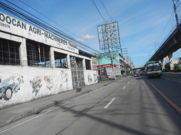 Stadtstra├če mit fahrenden Fahrzeugen, eine ├Überquerung auf der rechten Seite, Geb├Ąude, Strommasten mit Kabeln, bew├Âlktem Himmel und ein Geb├Ąude mit Graffiti an der Seite.