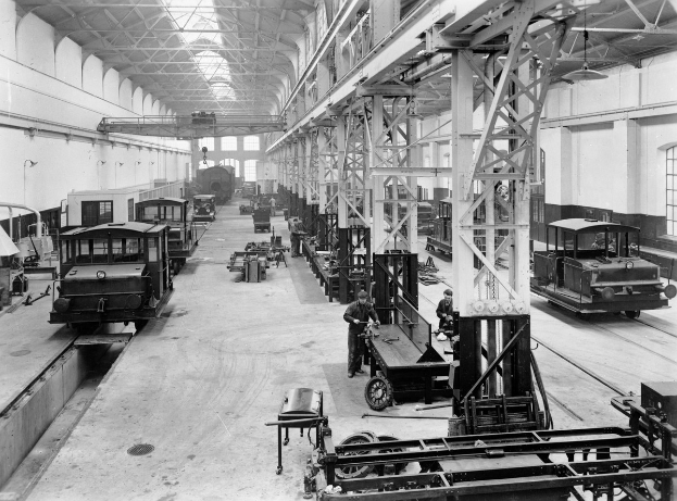 Schwarzes und weißes Foto einer Fabrikhalle mit mehreren Bahnen in Reparatur, Arbeitern, Metallstangen, Deckenlampen und Wandfenstern.