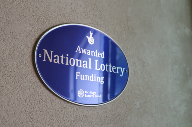 Blauer rechteckiger Schild mit weißem Hintergrund und schwarzer fettgedruckter Schrift, auf dem "Mit Nationaler Lotterie-Förderung ausgezeichnet" steht, an einem Gebäude angebracht.