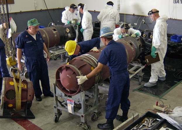 Mannergruppe in Helmen und Handschuhen beim Entschärfen einer großen Bombe in einer Fabrik mit Maschinen und Wandtext.