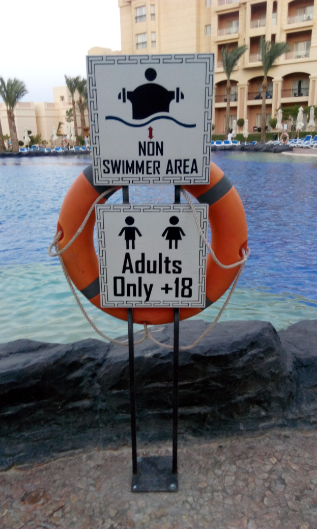 Ein Schwimmbad mit einem Schild "Nur für Erwachsene, die nicht schwimmen können" im Vordergrund, umgeben von Bäumen, Gebäuden und einem klaren blauen Himmel, mit einem Rettungsring in der Mitte des Pools.