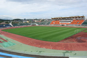 Großes Stadion mit einem Fußballfeld, umgeben von Gebäuden, Bäumen, Hügeln und einem klaren blauen Himmel, mit ein paar Menschen auf saftig grünem Gras.