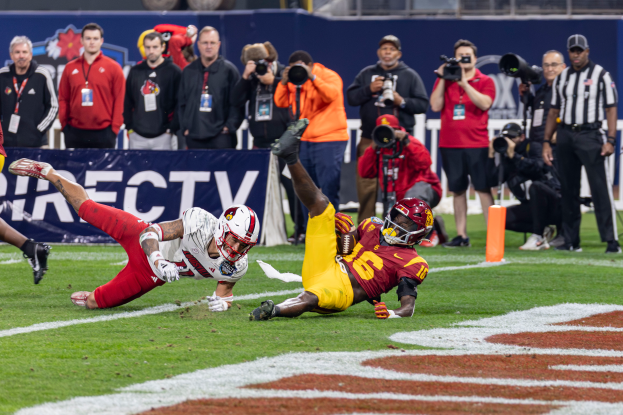 Ein Football-Spieler taucht in die Endzone ein, um einen Touchdown zu erzielen, mit einer Gruppe von Menschen am Boden, einige halten Kameras und ein Banner mit Text im Vordergrund, während Geländer, Pfosten und andere Gegenstände im Hintergrund sichtbar sind.