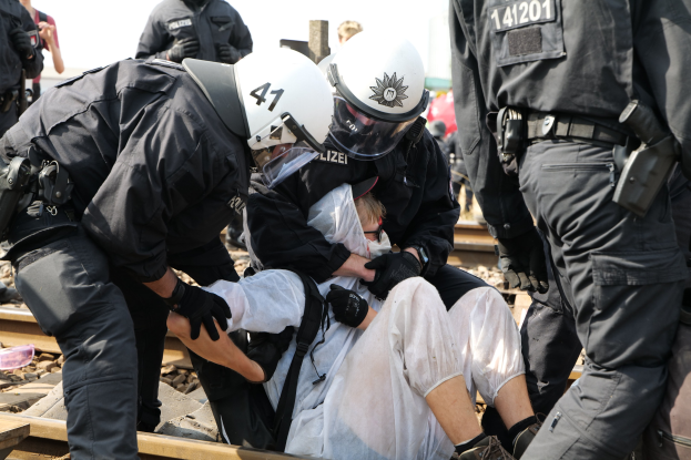 Eine Gruppe von Polizisten in Einsatzausrüstung nimmt einen Mann in einem weißen Kleid auf einem Gleis fest, andere sind im Hintergrund zu sehen.