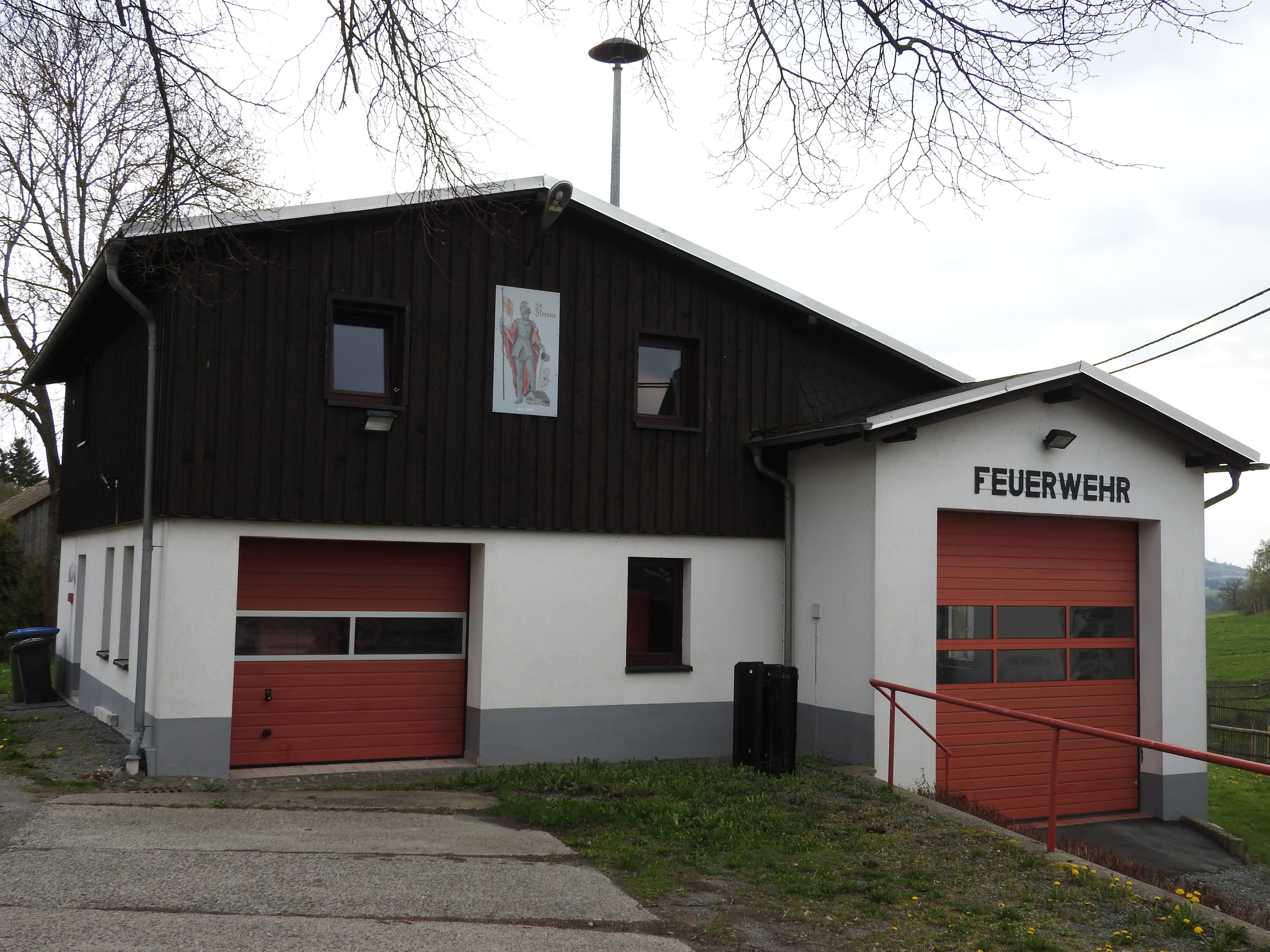Feuerwache mit rotem und weißem Gebäude, Straße, Gras, gelben Blumen und Bäumen vor einem Himmel als Hintergrund.