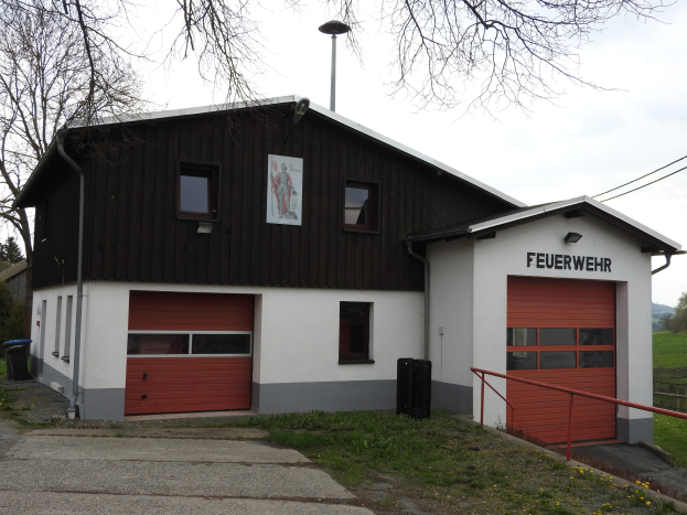 Feuerwache mit rotem und weißem Gebäude, Straße, Gras, gelben Blumen und Bäumen vor einem Himmel als Hintergrund.