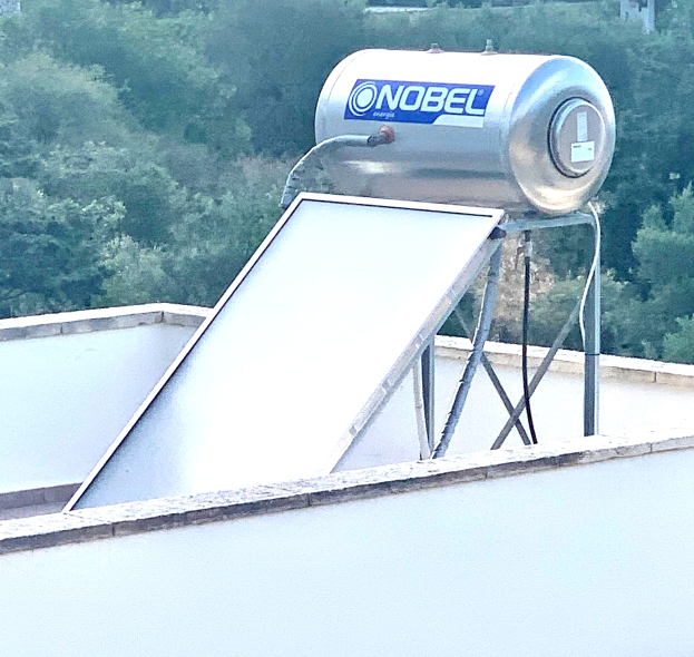 Solarwassererwärmer auf dem Dach eines Gebäudes mit einem Speicherbehälter oben, Bäume im Hintergrund.