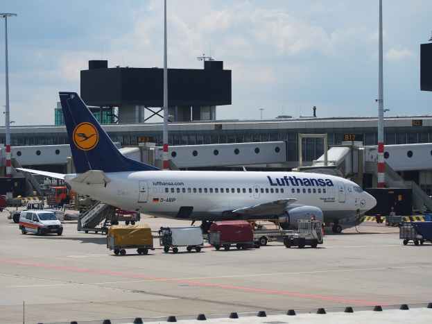 Lufthansa Airbus A320-200 Flugzeug am Frankfurt Airport geparkt mit Bodenfahrzeugen und Wagen im Vordergrund, ein Gebäude und Laternenpfähle im Hintergrund unter einem klaren blauen Himmel.