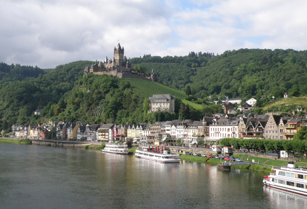 Ein idyllischer Blick auf den Rhein in Deutschland mit einer Burg auf einem Hügel, Booten auf dem Fluss und Fahrzeugen auf der Straße sowie einem bewölktem Himmel.