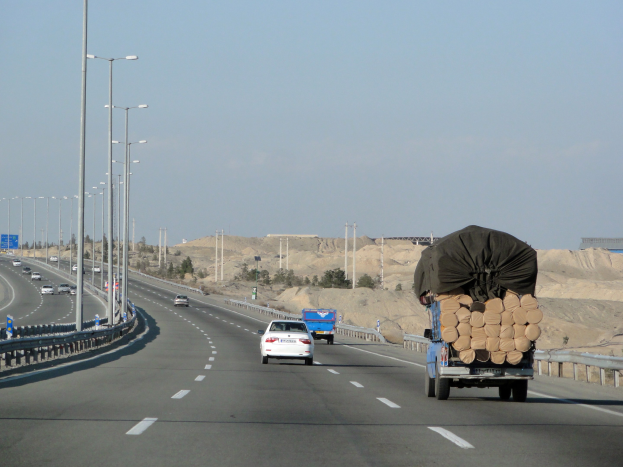 Ein Holzlaster mit einer großen Ladung Holz fährt auf einer Autobahn, umgeben von Geländern, Laternen, Schildern, Bäumen und Sand, mit Hügeln und einem klaren blauen Himmel im Hintergrund.