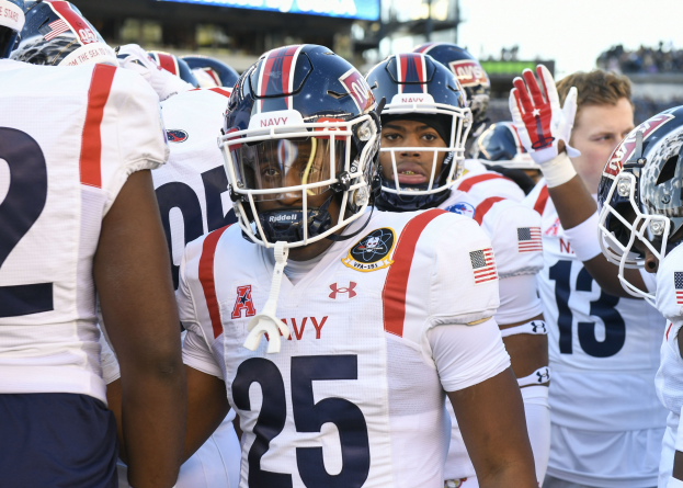 Gruppe von Männern in Fußballausrüstung auf einem Feld mit Helmen und Handschuhen, mit einer unscharfen Menge, Anzeigetafel mit 'Auburn vs Virginia Tech - 11/28/15 College Football Pick, Odds, and Prediction' und Himmel im Hintergrund.