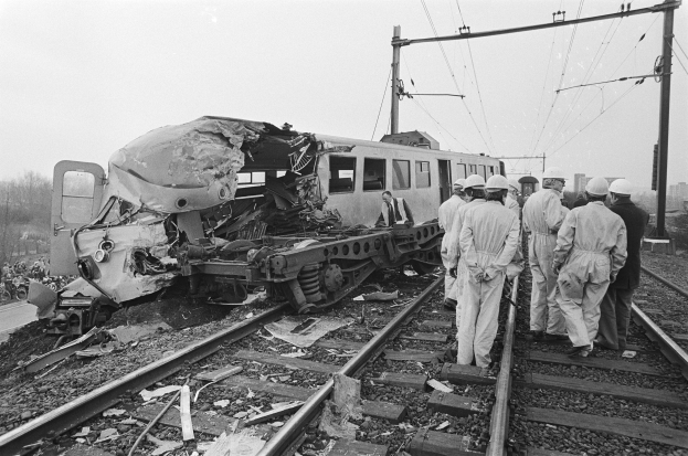 Schwarz-weiß-Bild eines Zugunglücks auf den Schienen mit Einsatzkräften in Helmen und einem Hintergrund von Strommasten, Bäumen, Gebäuden und einem klaren Himmel.