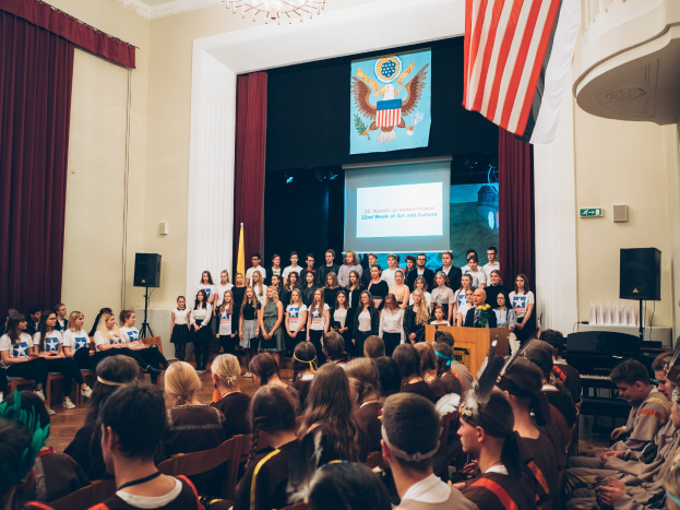 Eine Gruppe von Menschen steht an einem Podium mit einem Mikrofon während eines 2. Wochenende von Kunst und Kultur, flankiert von Fahnen und Lautsprechern, gegenüber einer sitzenden Zuschauermenge mit einem Bildschirm und einem Kronleuchter im Hintergrund.