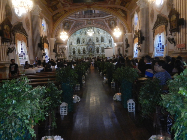 Eine Trauung in einer Kirche mit Gästen auf Bänken und im Mittelgang sitzend, dekoriert mit Zimmerpflanzen und Skulpturen, beleuchtet von Kronleuchtern und Fenstern mit Vorhängen und einem Bildschirm im Hintergrund.