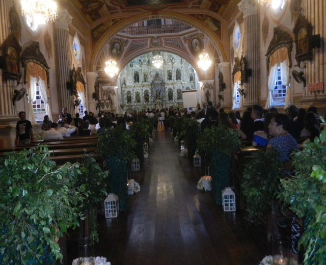 Eine Trauung in einer Kirche mit Gästen auf Bänken und im Mittelgang sitzend, dekoriert mit Zimmerpflanzen und Skulpturen, beleuchtet von Kronleuchtern und Fenstern mit Vorhängen und einem Bildschirm im Hintergrund.