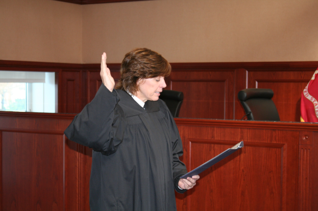 Eine Frau in einer schwarzen Robe steht vor einem Richtertisch, hält eine Akte, mit einem Mikrofon und einer Flagge auf dem Tisch, Stühle und ein Fenster im Hintergrund.