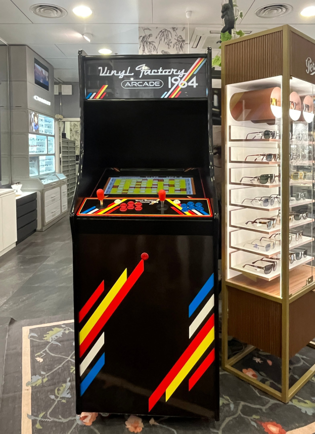 Ein Geschäft mit einem zentralen Retro-Arcade-Automaten, einem Regal mit Gläsern auf der rechten Seite und Hintergrundschränken, Fernseher und anderen Gegenständen, mit Deckenleuchten, die eine retro-mäßige Atmosphäre schaffen.