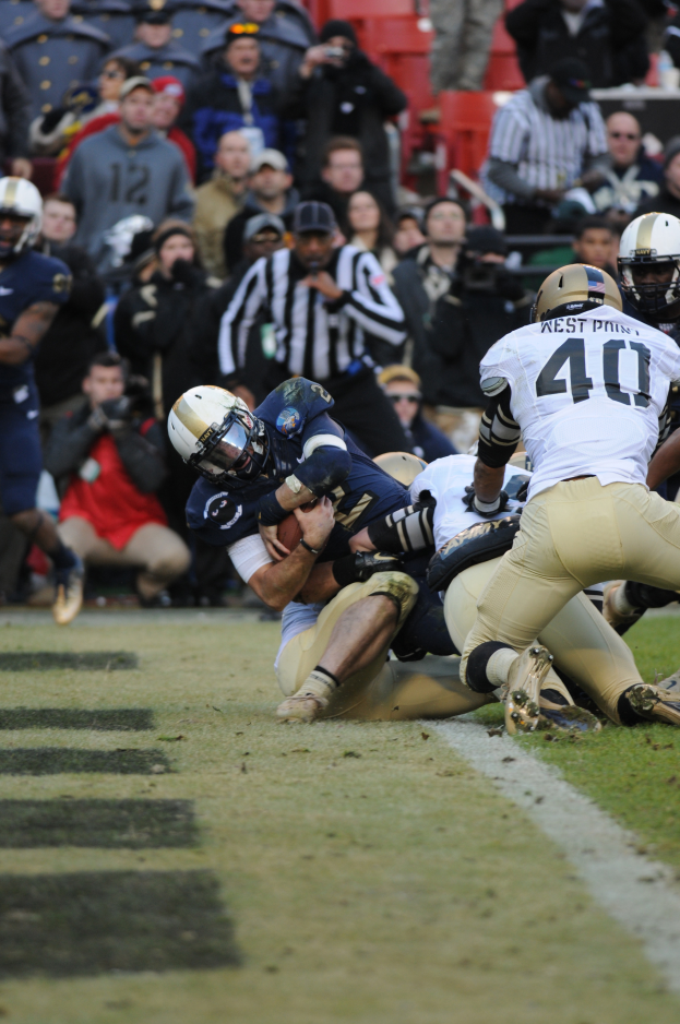 Ein Fussballspieler wird von einer Gruppe von Verteidigern auf einem Rasenfeld getackelt, mit Zuschauern und Kameramännern im Hintergrund, alle Spieler tragen Helme.