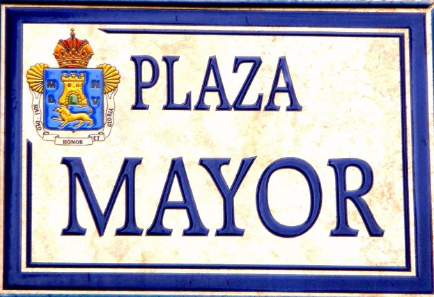 Rechtwinkliges weißes Schild mit schwarzer Schrift "Plaza Mayor" in fetter Schrift, blauem Rand und einem zentralen gekrönten Wappen-Logo an einer Gebäudefassade.
