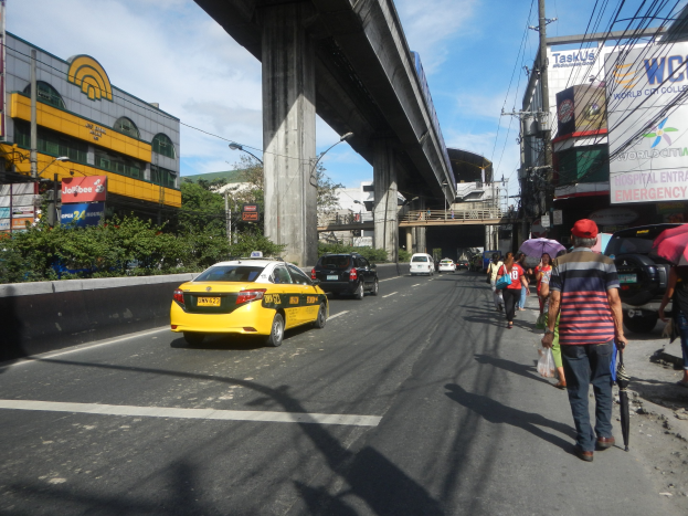 Ein gelbes Taxi fährt eine Straße mit hohen Gebäuden entlang, wo Fußgänger mit Regenschirmen gehen, eine Brücke ist im Hintergrund zu sehen und Stromleitungen verlaufen über die Straße unter einem bewölkten Himmel.