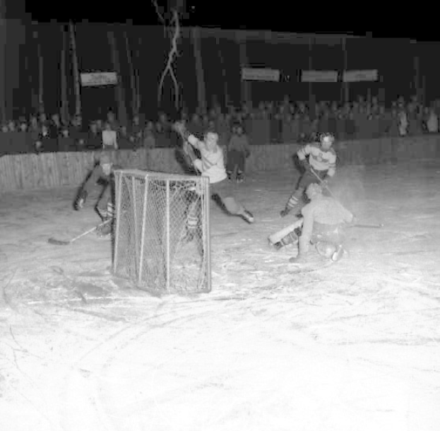 Schwarzes und weißes Foto eines Hockey-Spiels auf einem Eisstadion, mit Spielern in der Nähe eines Tores und Zuschauern im Hintergrund.