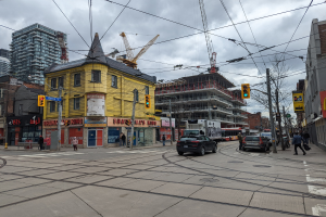 Ein gelbes Gebäude im Bau auf einer Stadtstraße mit Autos, Fußgängern, Ampeln, Strommasten, Kränen und einem bewölkten Himmel.