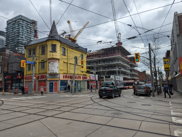 Ein gelbes Gebäude im Bau auf einer Stadtstraße mit Autos, Fußgängern, Ampeln, Strommasten, Kränen und einem bewölkten Himmel.