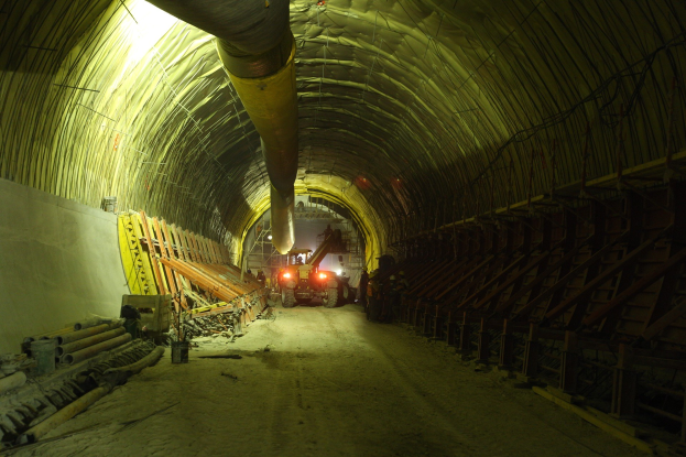 Baustelle mit einem mittigen Tunnel, Fahrzeugen, verstreuten Holzobjekten, Rohren und einer linken Wand, mit Überkopf- und Hintergrundbeleuchtung.