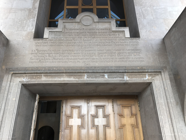 Große hölzerne Tür vor einem steinernen Gebäude mit einem Glasfenster oben, mit Inschrift auf der Wand, die wahrscheinlich Holocaust-Opfer gedenkt.