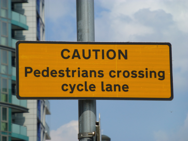 Gelbes Warnschild an einem Pfahl vor einem Gebäude, auf dem steht: "Fußgänger überqueren Fahrradspur" mit einem klaren blauen Himmel im Hintergrund.
