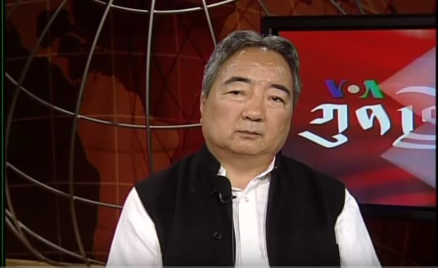 Ein Mann in schwarzer Weste und weißem Hemd sitzt vor einem Fernsehbildschirm und spricht in ein Mikrofon, im Hintergrund ist eine Weltkugel zu sehen.