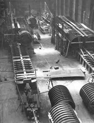 Schwarzes und weißes Foto eines industriellen Fabrikinneren mit zahlreichen Rohren, Maschinen und Geräten auf dem Boden, vor strukturellen Säulen und einer Wand.
