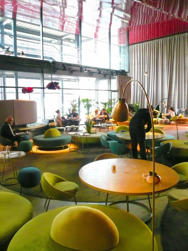 Ein geräumiges Hotel-Lounge mit grünen Stühlen, Tischen, Sofas, stehenden Menschen, Pflanzen, einer Stehlampe, Vorhängen, Deckenleuchten und einer gemütlichen Atmosphäre.