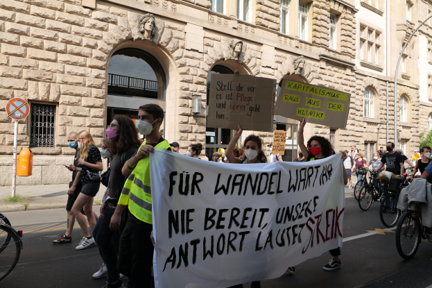 Eine Gruppe von Menschen marschiert auf der Straße in Berlin, einige halten Schilder und Banner, andere fahren Fahrräder, mit einem Gebäude im Hintergrund, das Fenster, Bögen, Säulen, Skulpturen und Bäume aufweist.