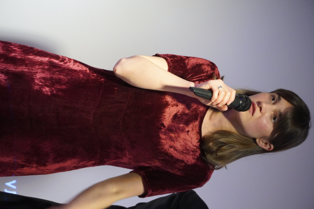 Eine Frau in einem roten Kleid steht selbstbewusst auf einer Bühne, hält ein Mikrofon in der Hand und hat eine glatte Bob-Frisur und natürliches Make-up.