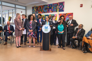 Gruppe von Menschen vor einem Podium mit Mikrofon und einer Tafel mit der Aufschrift "Sancho-Stadtrat tritt dem Stadtrat bei", mit sitzenden und stehenden Teilnehmern, Flaggen, Feuerlöscher und Deckenleuchten im Hintergrund.