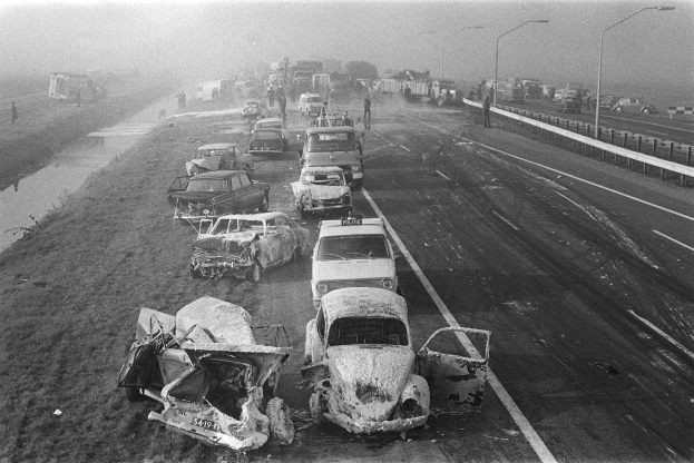 Ein Schwarz-Weiß-Foto von mehreren verunglüten Autos am Straßenrand bei Nacht, mit Laternen, nahestehenden Menschen, Bäumen und einem Gewässer im Hintergrund.