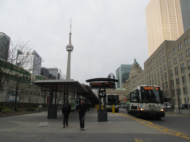 Eine belebte Stadtstraße mit einem Bus an einer Bushaltestelle, Passanten auf dem Gehweg, Fahrzeuge auf der Straße, ein kleines Häuschen, Bäume, hohe Gebäude, ein Turm und ein klarer Himmel.