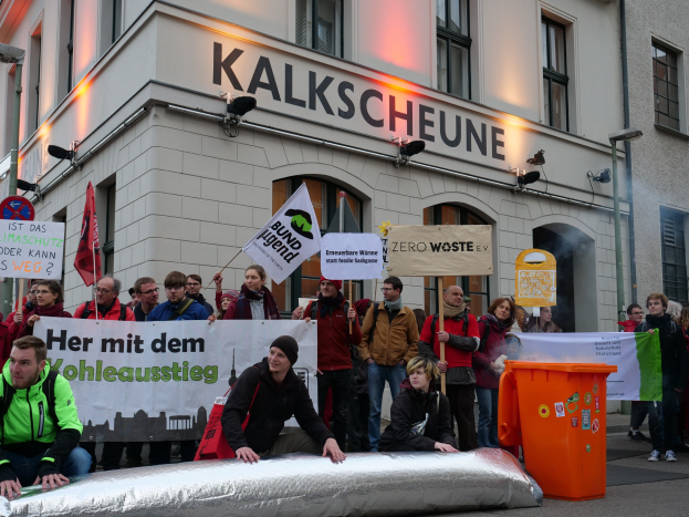 Eine Gruppe von Menschen mit Schildern und Plakaten vor einem Gebäude bei einer Demonstration in Deutschland, mit zwei Personen auf einem Objekt sitzend im Vordergrund und einem Müllbehälter auf der rechten Seite.