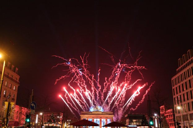 Eine belebte Stadtstraße am Silvesterabend in Berlin, voller Menschen, Fahrzeuge und Gebäude, erhellt von Lichtern und Feuerwerk am Himmel.