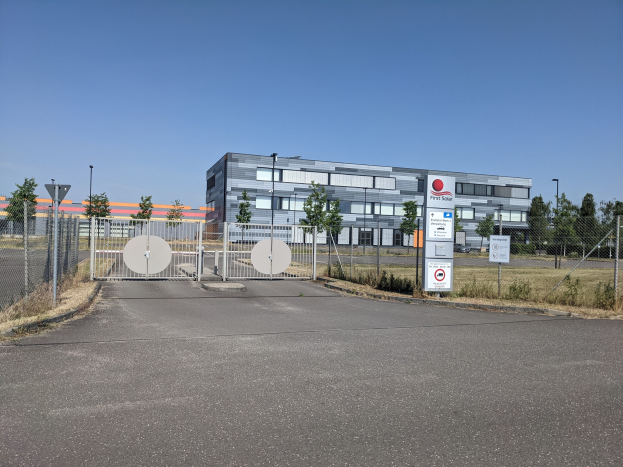 Großes industrielles Gebäude mit der Aufschrift "First Solar" und vielen Fenstern, umgeben von einem Metallzaun, umgeben von Bäumen, Pflanzen, Gras, Schildern, Straßenlaternen und einem Weg unter einem bewölkten Himmel.