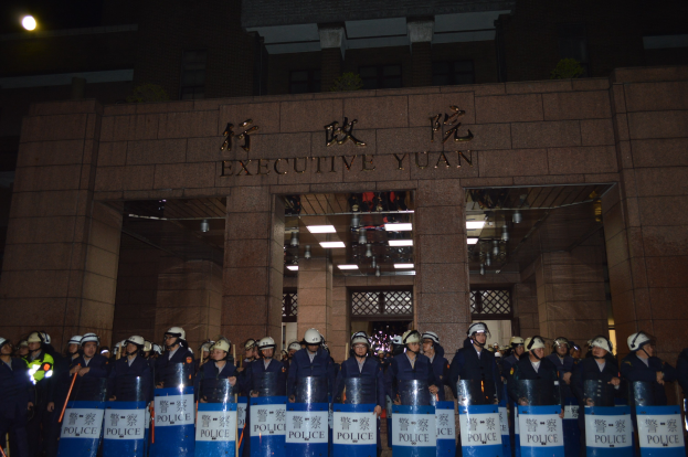 Gruppe von Polizisten in Helmen und Schilden vor einem Geb├Ąude mit beleuchteter Wand- und Deckenbeschriftung, mit weiteren Geb├Ąuden im Hintergrund.