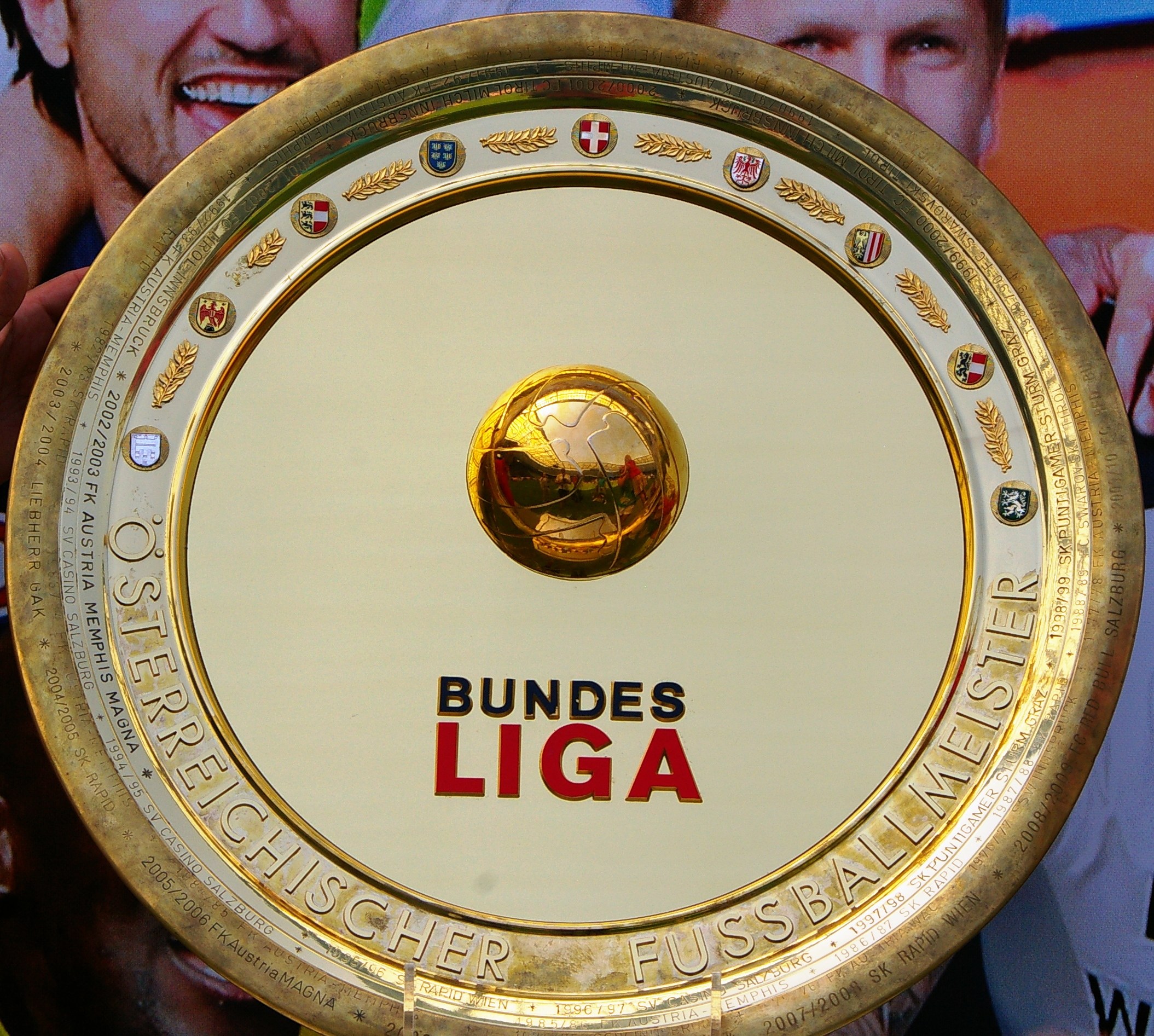 Goldener Schild mit 'Bundesliga'-Text in den Händen eines Mannes, mit einem Banner im Hintergrund mit drei Personen.