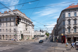 Eine Stadtstraße mit Autos und Fußgängern, umgeben von Gebäuden, Strommasten, Verkehrszeichen, Schildern, Bäumen und einem bewölkten Himmel.