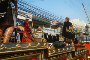 Eine Gruppe von Menschen in Kostümen fährt auf einem hell erleuchteten Umzugswagen, mit Gebäuden, Schildern, Pfählen, Drähten, Bäumen und einem klaren blauen Himmel im Hintergrund.