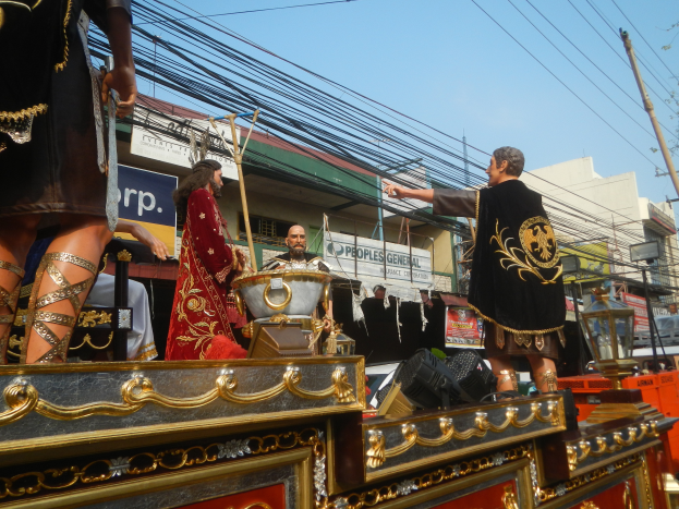 Eine Gruppe von Menschen in Kostümen fährt auf einem hell erleuchteten Umzugswagen, mit Gebäuden, Schildern, Pfählen, Drähten, Bäumen und einem klaren blauen Himmel im Hintergrund.