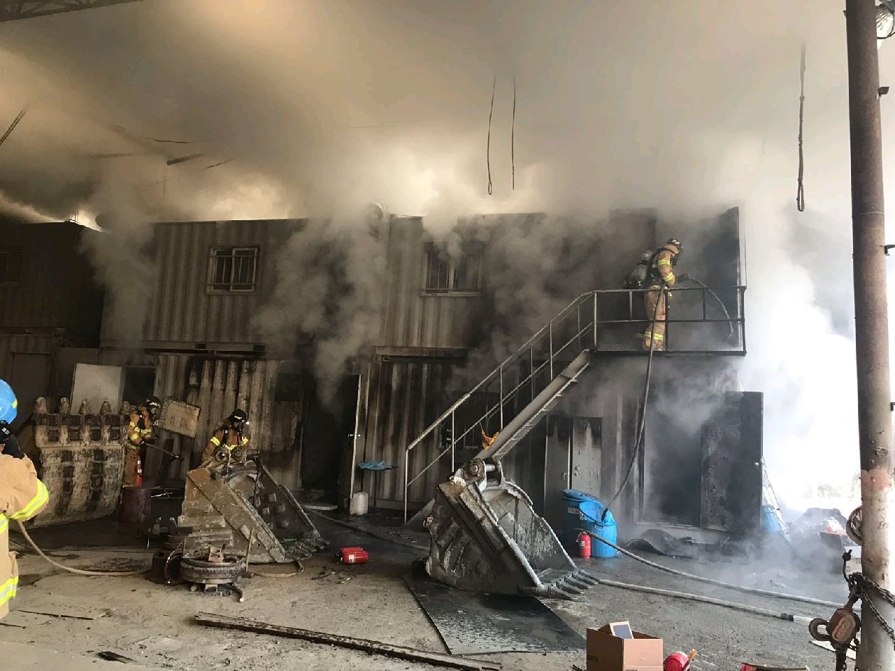 Feuerwehrleute in Helmen verwenden Schläuche, um ein Lagerfeuer zu löschen, mit Rauch, der aus dem Gebäude aufsteigt und Trümmern, die draußen verstreut sind.