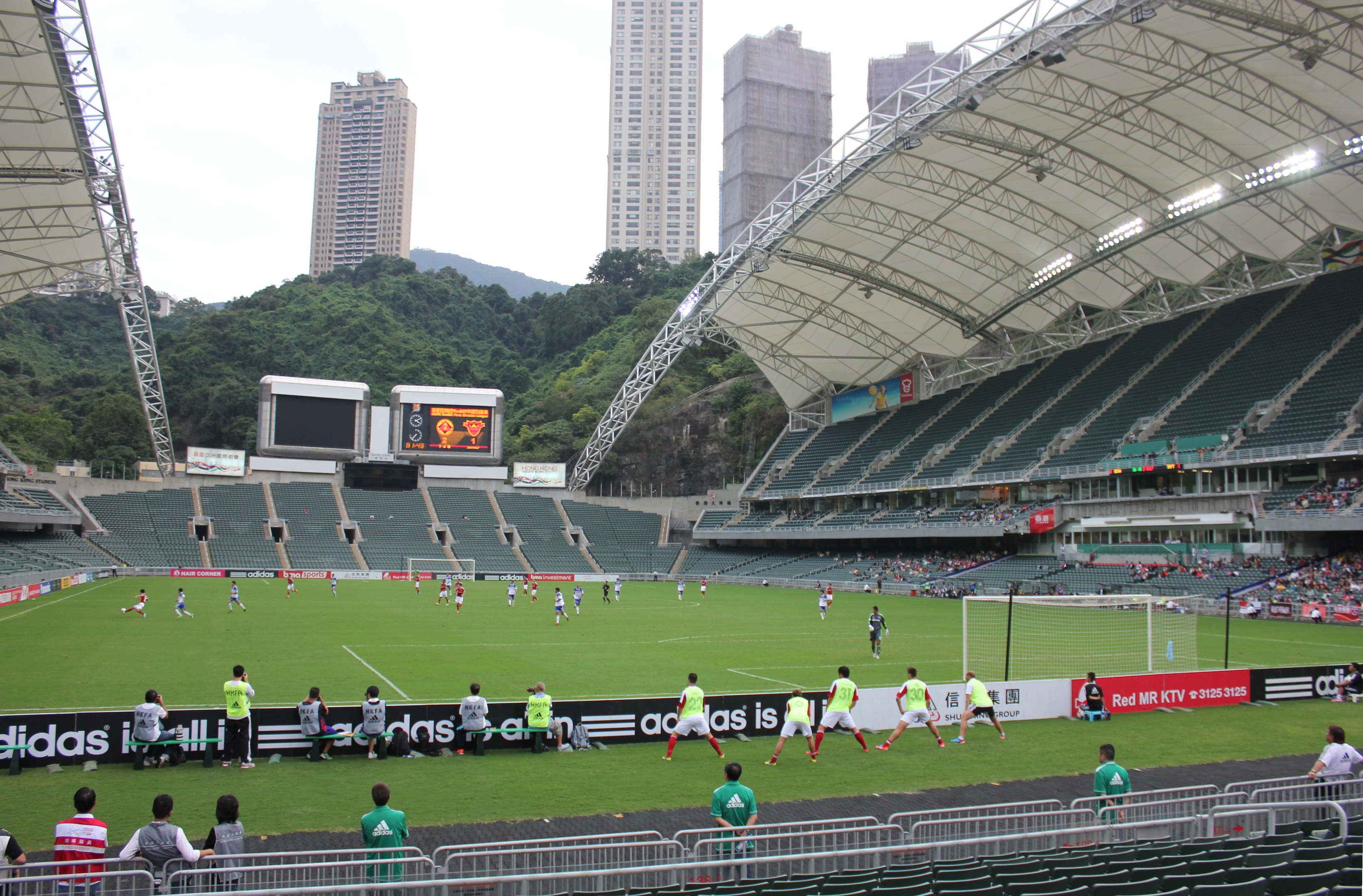 Ein Fußballspiel in einem großen Stadion mit Zuschauern, Bannern, einem Tor, Infrastruktur und einer bewölkten Himmelslandschaft.