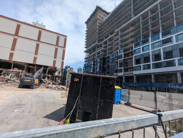 Großes Gebäude mit Schutt davor, umgeben von einem Metallzaun, einem Container, Pflanzen, Bäumen, Pfählen, einem Schild, Fahrzeugen, Steinen und einem bewölkten Himmel an einer Baustelle für ein neues Wohngebäude.
